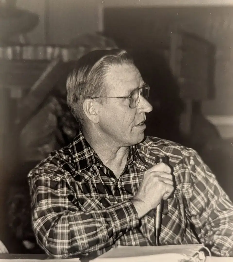 Black-and-white photo of an older man in glasses and a plaid shirt, holding a microphone and looking to his left. Papers are visible in front of him.