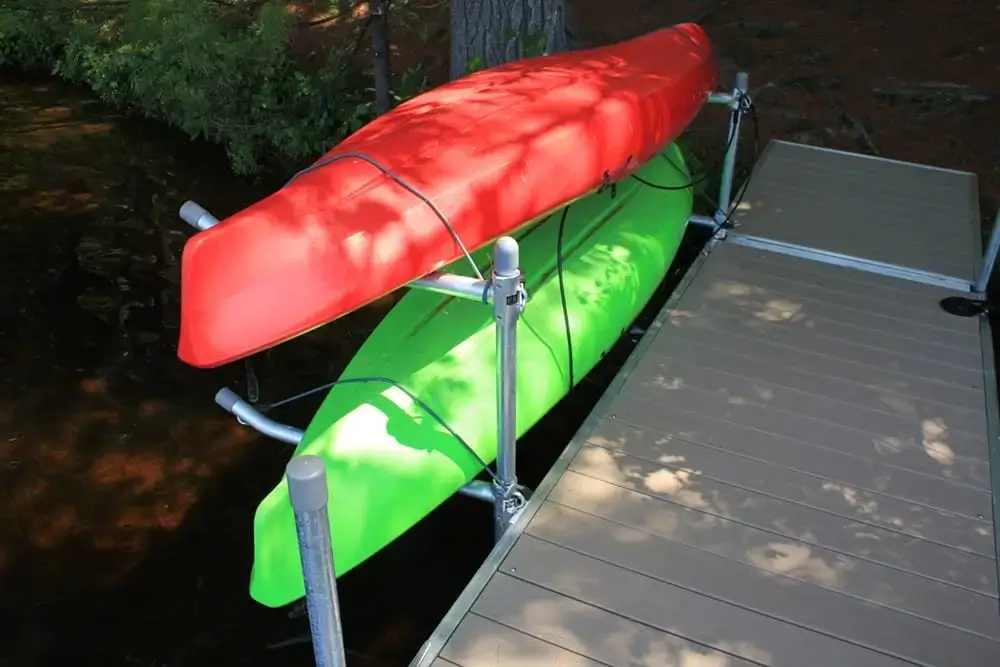 Two kayaks, one red and one green, are stored on a metal rack next to a wooden dock by the water, with trees and foliage in the background.