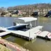 A Connect-A-Dock with a covered seating area and red chairs floats on a calm lake, with houses and trees in the background.
