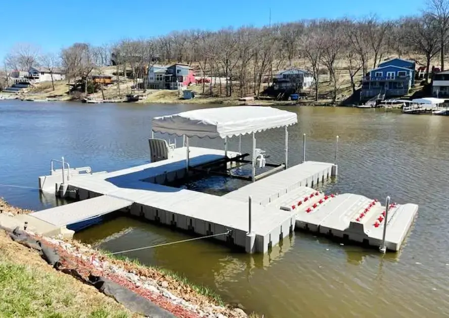 IMG_20230412_152218 A Connect-A-Dock with a covered seating area and red chairs floats on a calm lake, with houses and trees in the background.
