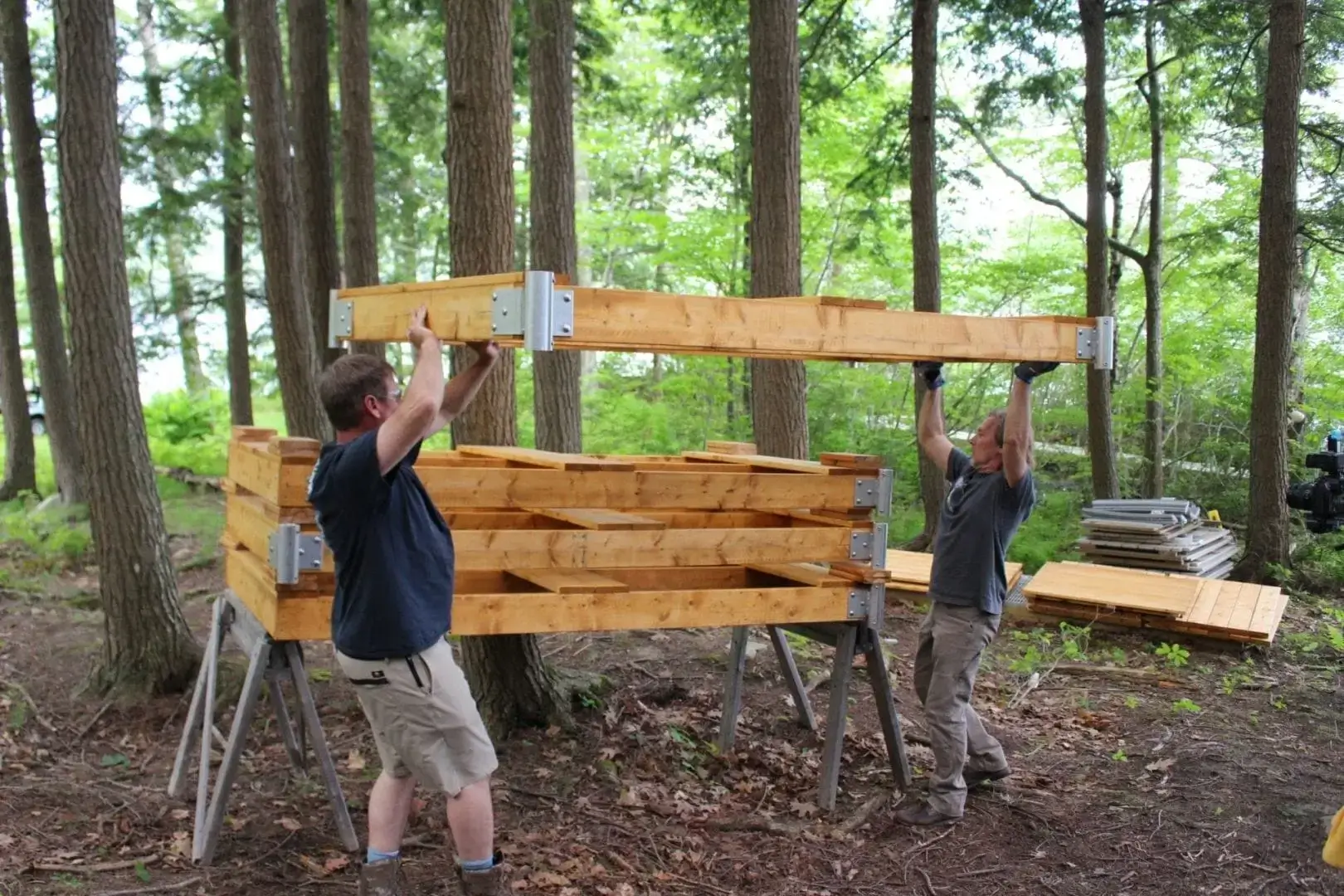 Two people lift a large wooden frame together in a forested area, assembling a wooden structure on metal sawhorses among tall trees.