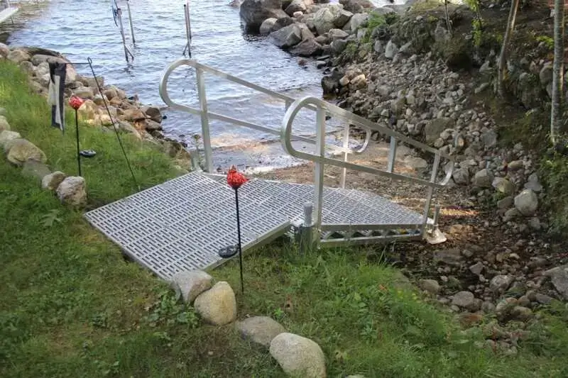 A metal wheelchair accessible ramp with handrails leads down to docks along a rocky shoreline, next to a grassy area bordered by stones.