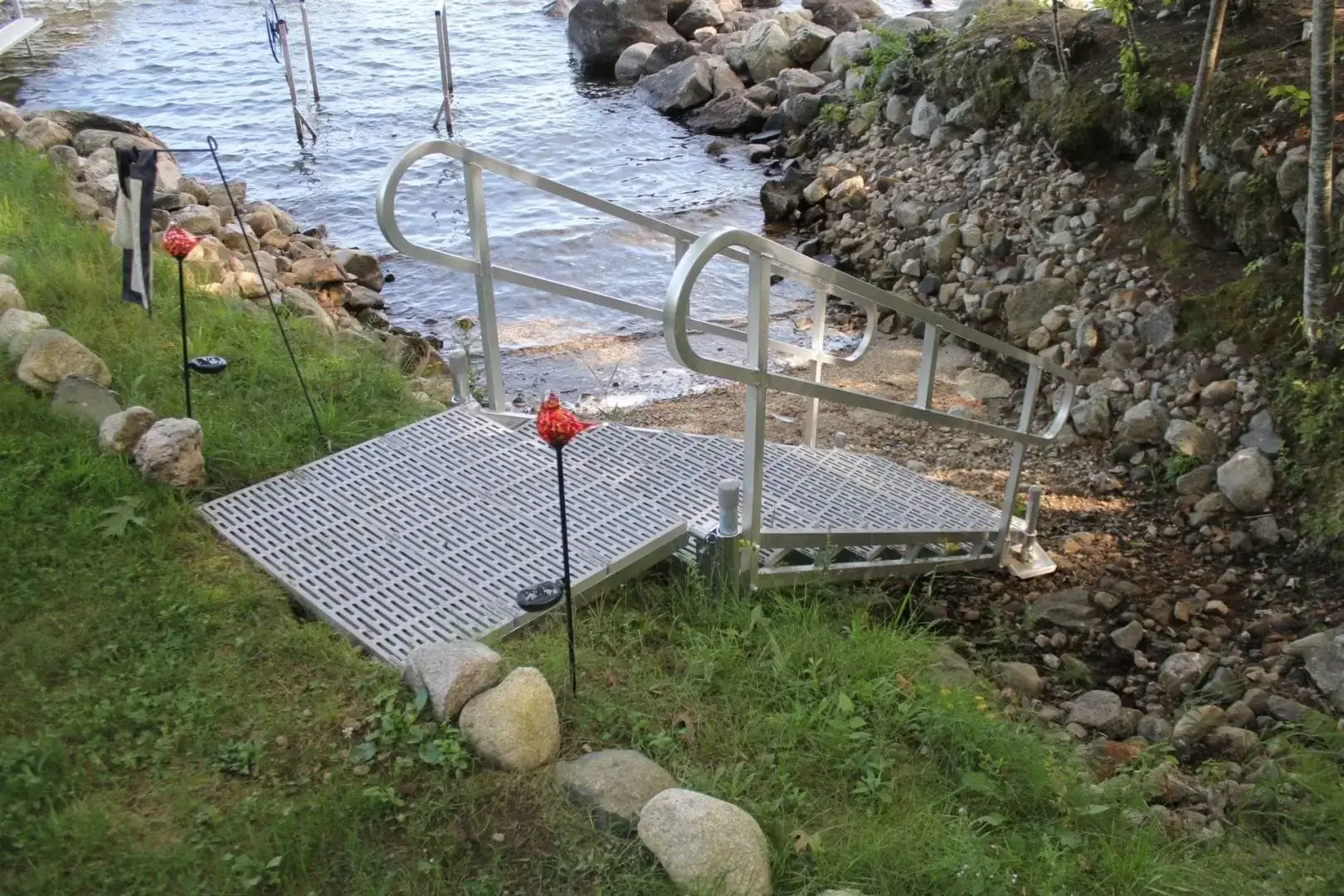 A metal ramp with railings leads from a grassy area down to a rocky shore by a lake, surrounded by stones and decorated with small torch lights.