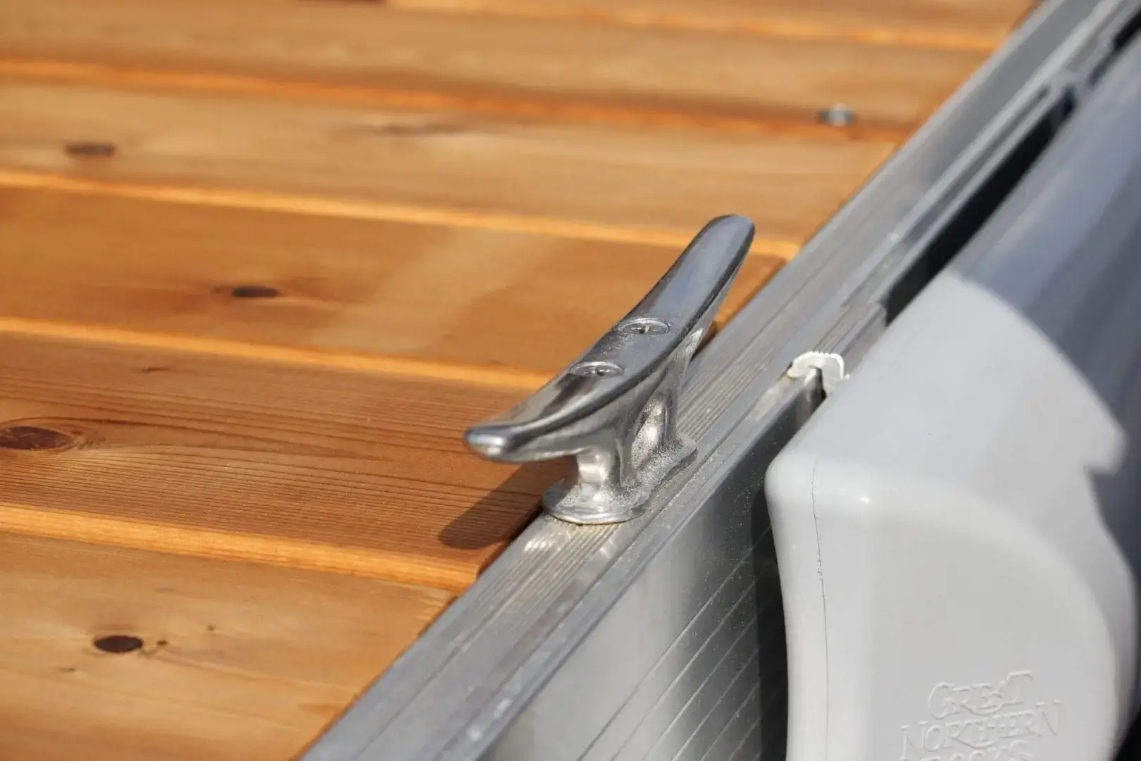A close-up of a metal boat cleat attached to the edge of a wooden dock with light brown planks.