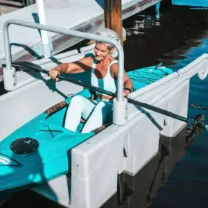 An athlete sits in a blue kayak on the water, preparing to launch using the Connect-A-Dock Kayak Launch system while holding a paddle.