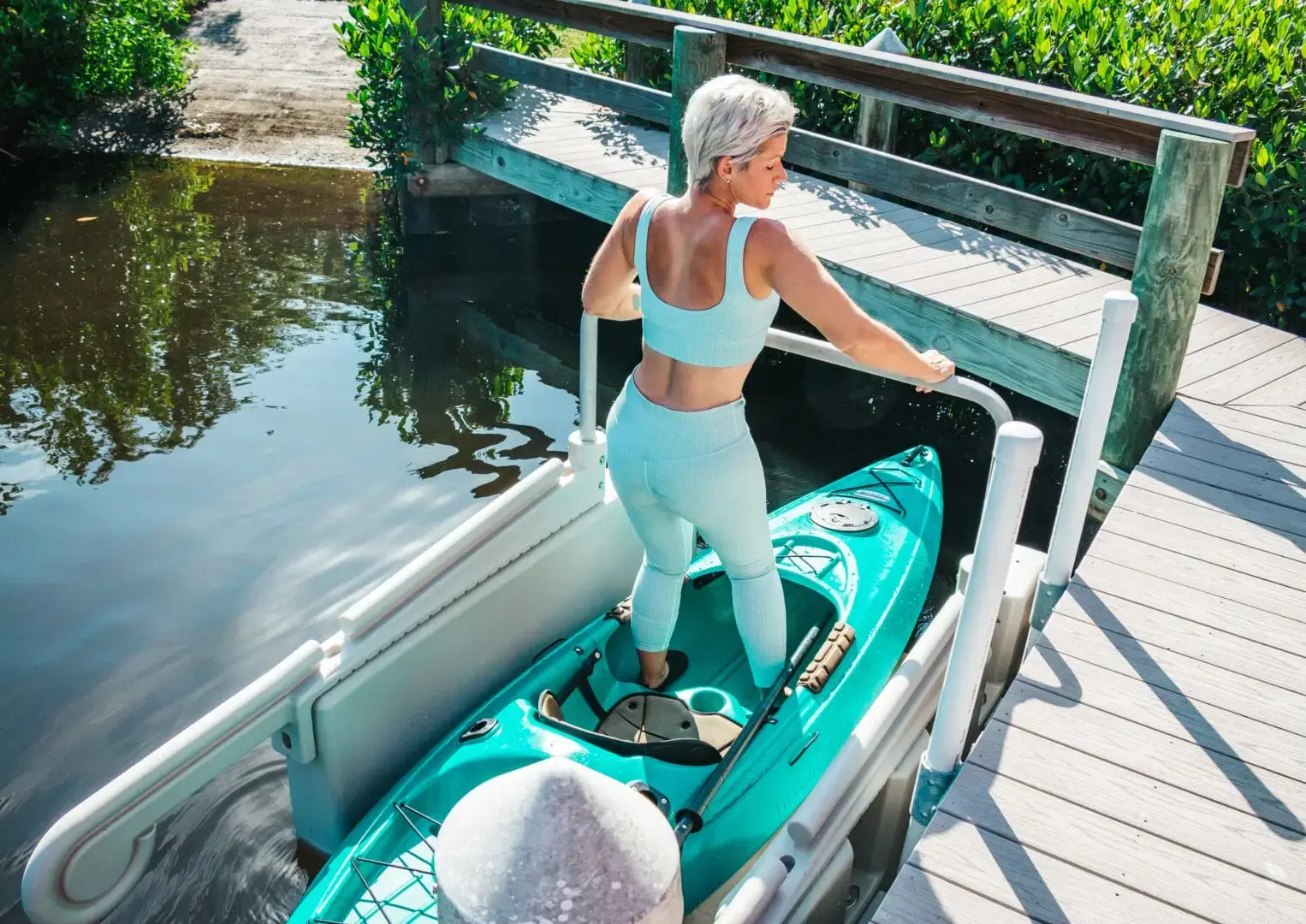 IMG_6998 A woman in light blue activewear readies a teal kayak on the Kayak Launch - Connect-A-Dock, surrounded by calm water and greenery.