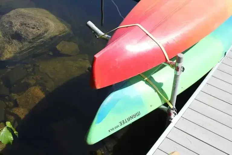 Two canoes, one red and one green, are stacked and tied together on docks beside clear water with visible rocks beneath the surface.
