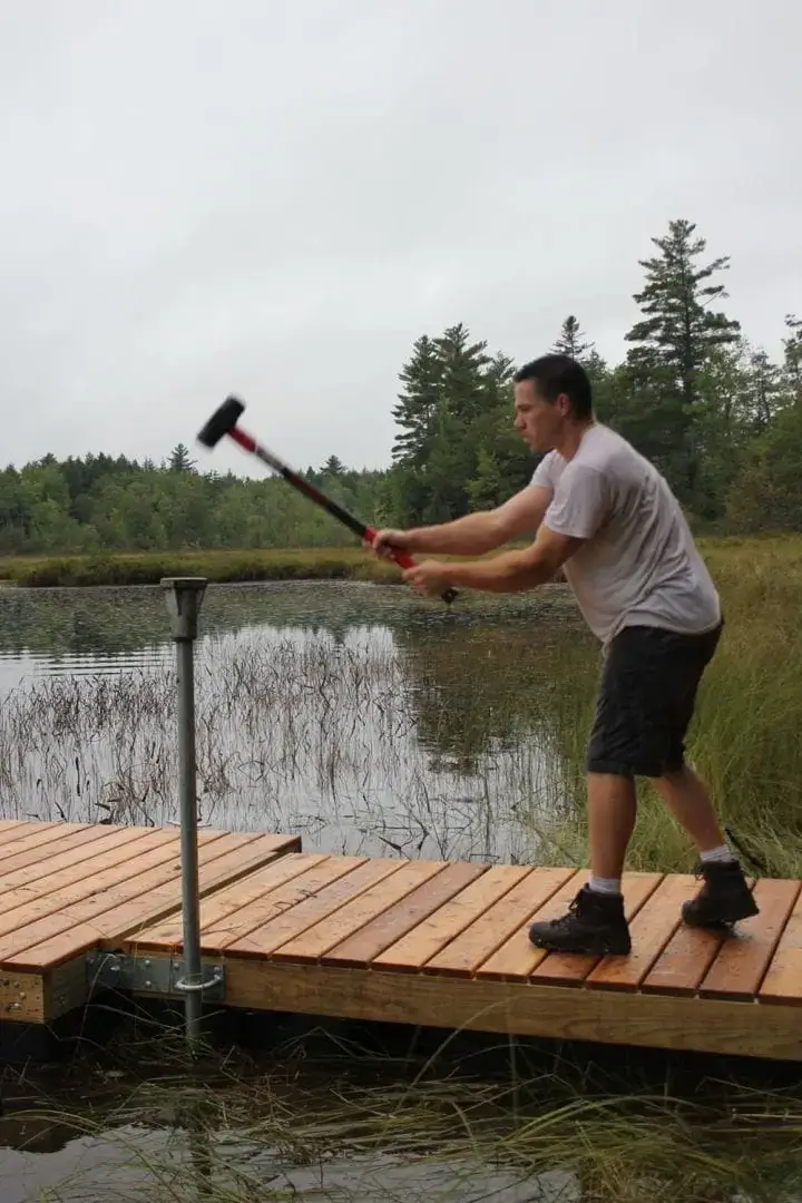 A man in a gray shirt and shorts uses a sledgehammer on a metal post by the edge of a wooden dock over a lake, surrounded by grassy vegetation and trees.
