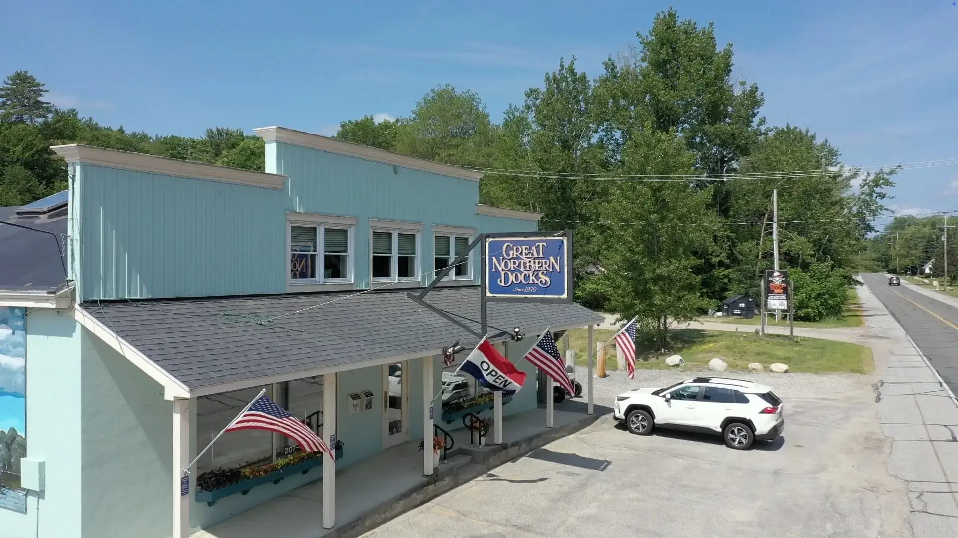 Light blue building with a “Great Northern Docks” sign, American flags, and an “OPEN” flag. A white SUV is parked outside, with trees and a road in the background.