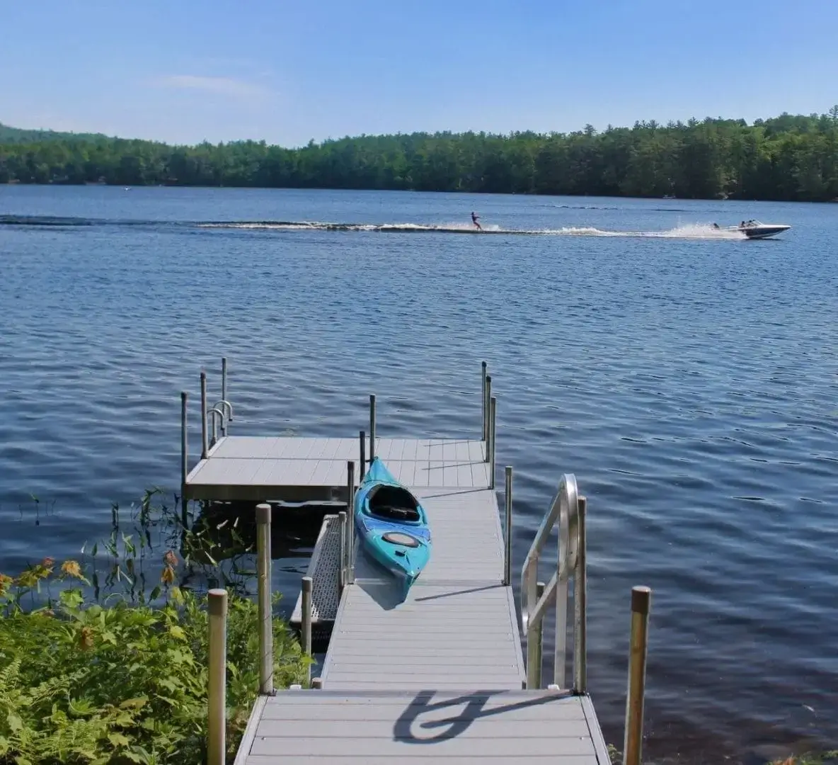 A blue kayak sits on a lakeside dock with calm water; a speedboat pulls a water-skier in the distance under a clear blue sky.