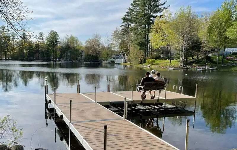 Two people sit on a bench at the end of a dock, enjoying a sunny day by the calm lake, surrounded by trees, houses, and other peaceful docks.