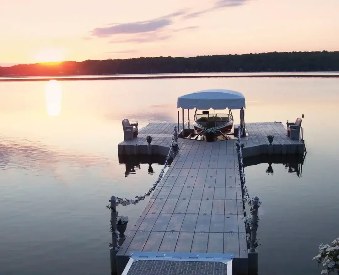 A Connect-A-Dock extends over calm water at sunset, featuring a covered boat, chairs, and flower pots, with serene views of the distant treeline.