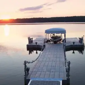 A Connect-A-Dock extends over calm water at sunset, featuring a covered boat, chairs, and flower pots, with serene views of the distant treeline.