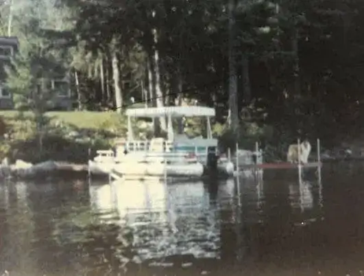 A white pontoon boat is docked by a wooden pier on a calm lake, surrounded by trees and a house partially visible in the background.
