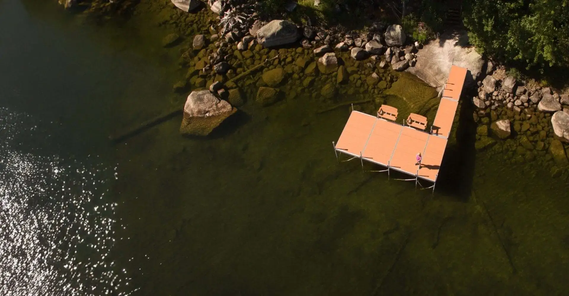 Aerial view of docks with lounge chairs extending over clear, shallow water and rocks by a forested shoreline.