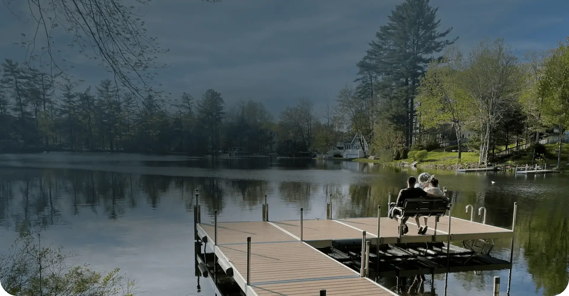 Two people sit on a bench at the end of a wooden dock overlooking a calm lake, surrounded by trees and houses, under a cloudy sky.