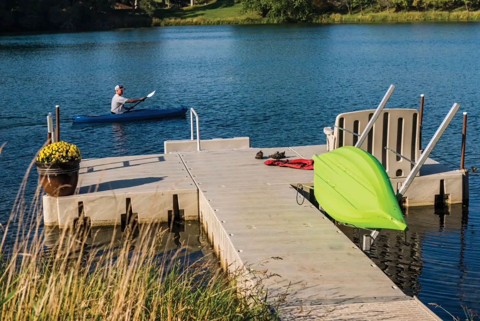 kayak_rack-03-low A man paddles a blue kayak on a lake near a Connect-A-Dock, which holds a green kayak, shoes, a red life vest, and a yellow-flower planter.