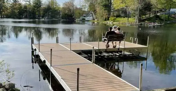 Two people sit together on a bench at the end of a dock overlooking a calm lake, surrounded by trees and houses on a sunny day.