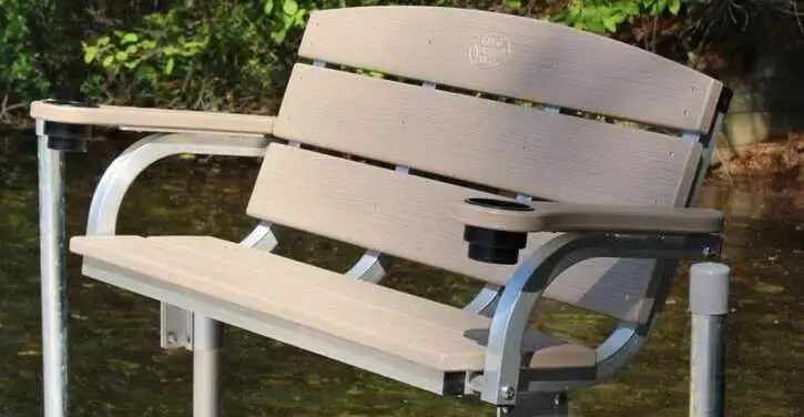 A light brown bench with metal armrests and built-in cup holders, positioned outdoors near water with greenery in the background.