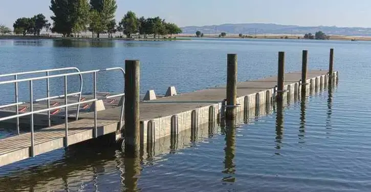 A wooden dock with railings extends over calm water, surrounded by trees and distant hills under a clear sky.