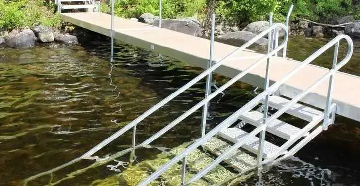 A dock extends over clear water with metal stairs and handrails leading down into the lake, surrounded by rocks and greenery.