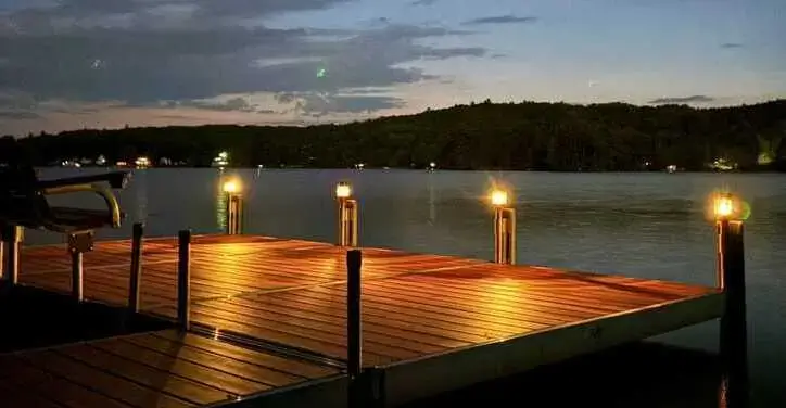 A wooden dock with glowing lights extends over a calm lake at dusk, with trees and a darkening sky in the background.