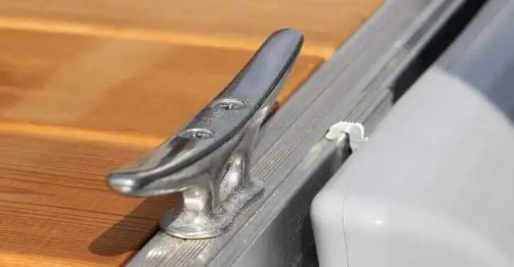 A metal boat cleat attached to the edge of a wooden dock, next to a gray inflatable boat.