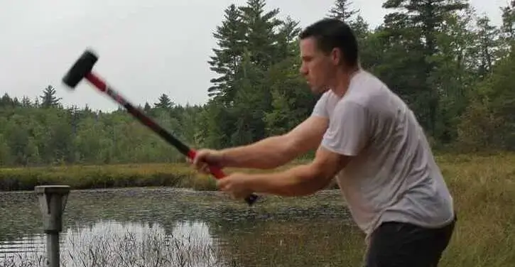 A man swings a sledgehammer outdoors near a body of water, surrounded by trees and greenery.