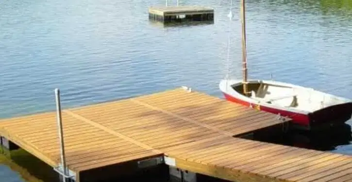A wooden dock extends over calm water, with a small white and red sailboat tied to it. A floating platform is visible in the distance.