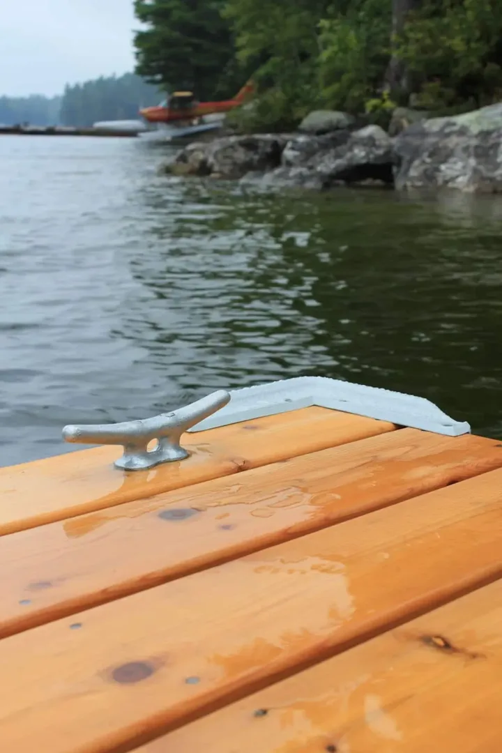 Close-up of a dock cleat on wet wooden planks at the edge of a lake, with rocky shoreline and trees visible in the background.