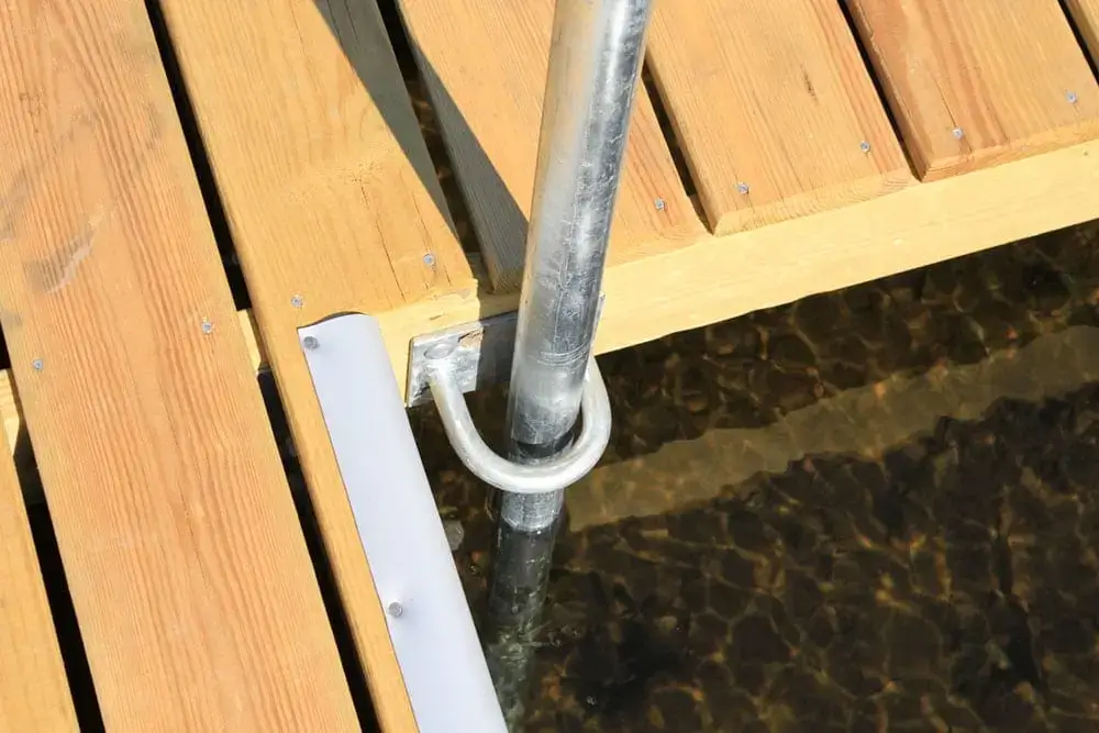 Close-up of a metal pole attached to the corner of a wooden dock above clear water, with visible wood grain and screws.