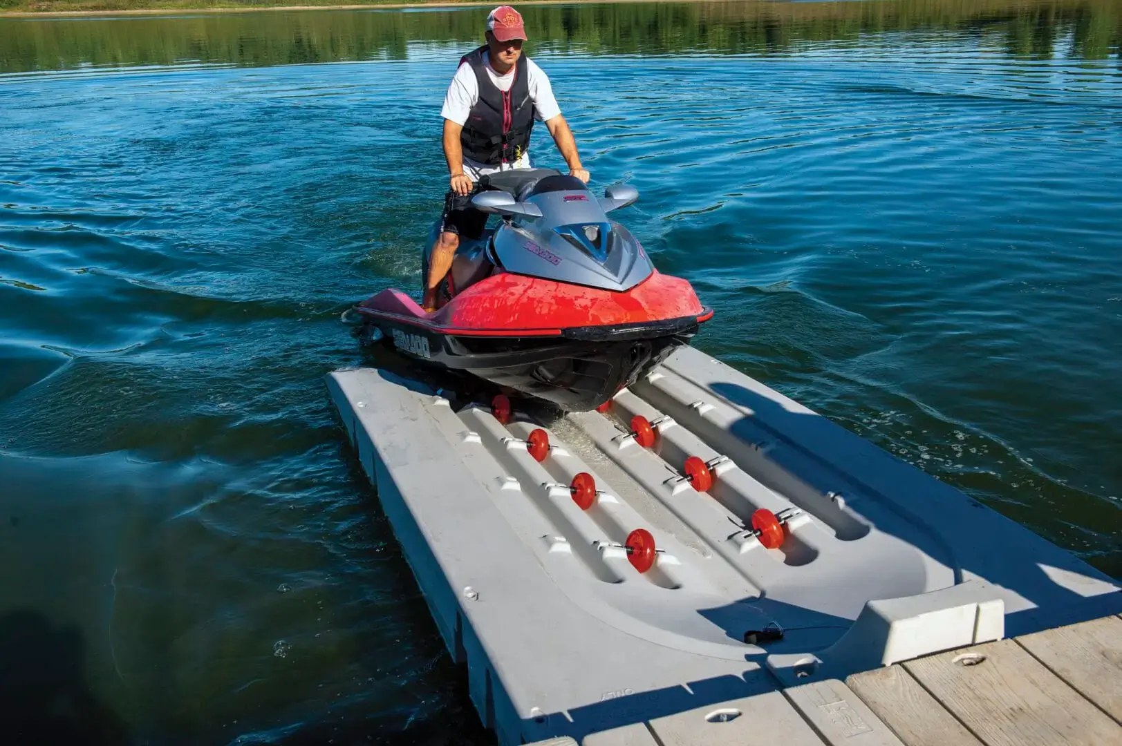 A person rides a red jet ski onto a PWC Port - Connect-A-Dock, surrounded by calm water and greenery.
