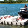 A person rides a jet ski onto a PWC Port - Connect-A-Dock floating dock on a sunny lakeside day, with trees and grassy shore in the background.