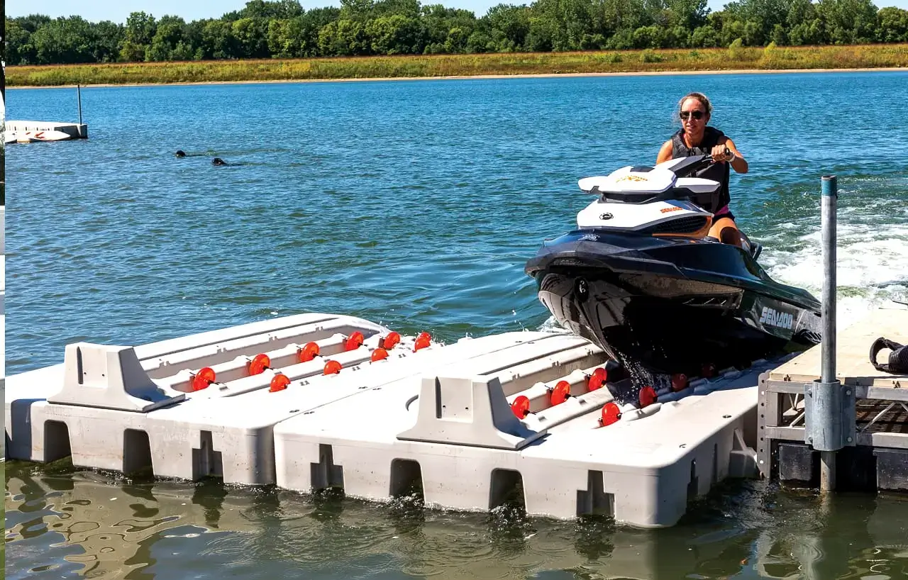 A person rides a jet ski onto a PWC Port - Connect-A-Dock floating dock on a sunny lakeside day, with trees and grassy shore in the background.