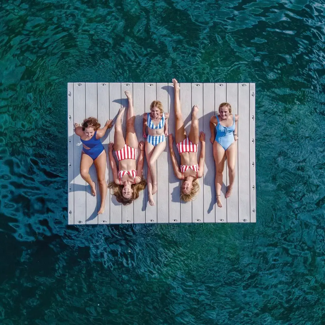 swim_platform-low Five women in swimsuits relax on a Swim Raft - Connect-A-Dock, floating on clear blue-green water, seen from above.