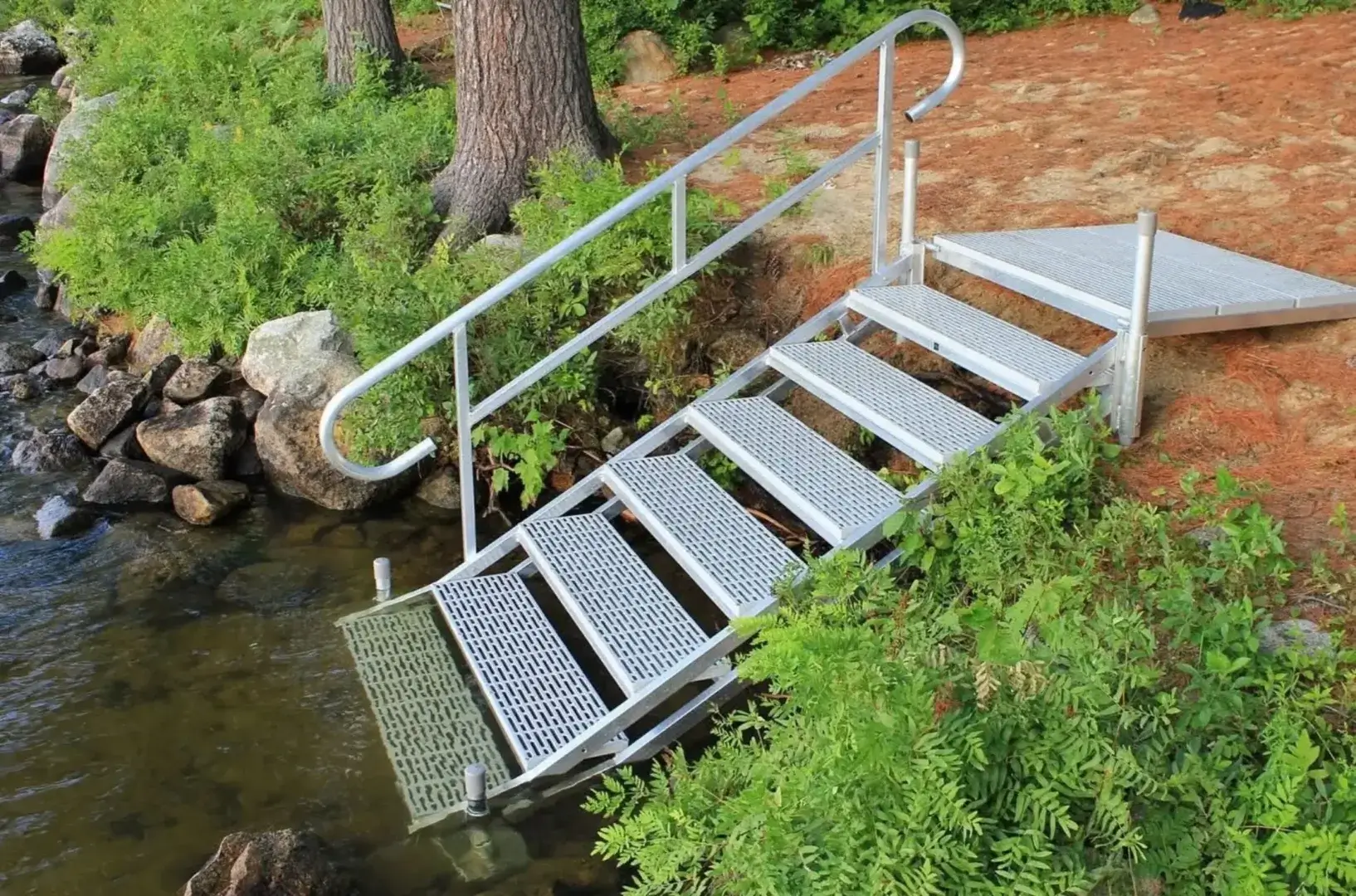 Metal stairs with a handrail lead from a forested, dirt-covered area down into a body of water, surrounded by green plants and rocks.