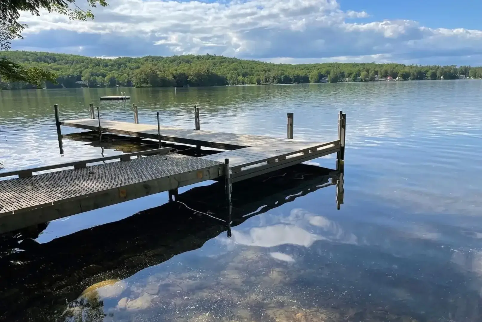 A wooden dock extends over a calm lake with clear water, surrounded by green trees and distant hills under a partly cloudy blue sky.
