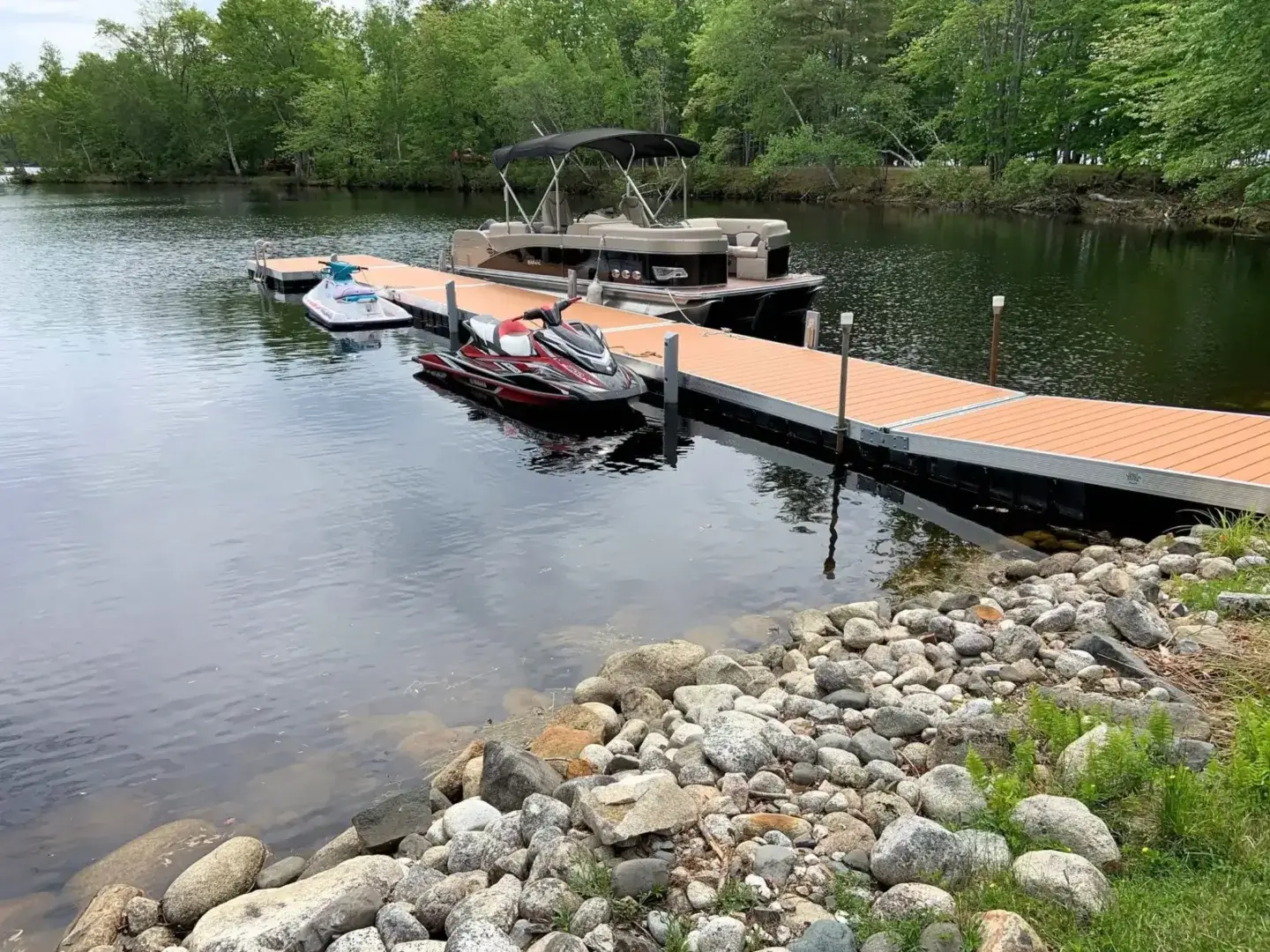 A wooden dock extends into a calm lake with jet skis and a pontoon boat moored; rocks line the shore, and trees surround the water.