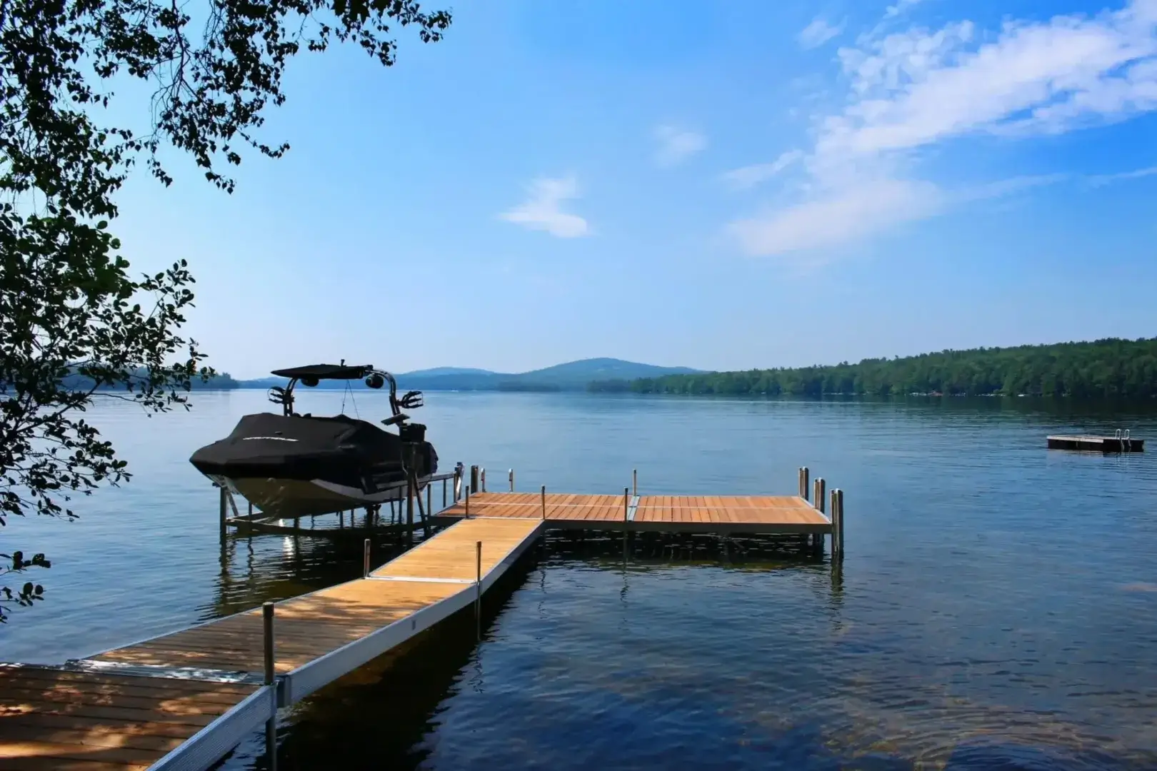 A boat is docked at a wooden pier on a calm lake with distant hills and a partly cloudy blue sky. Trees frame the waters edge.