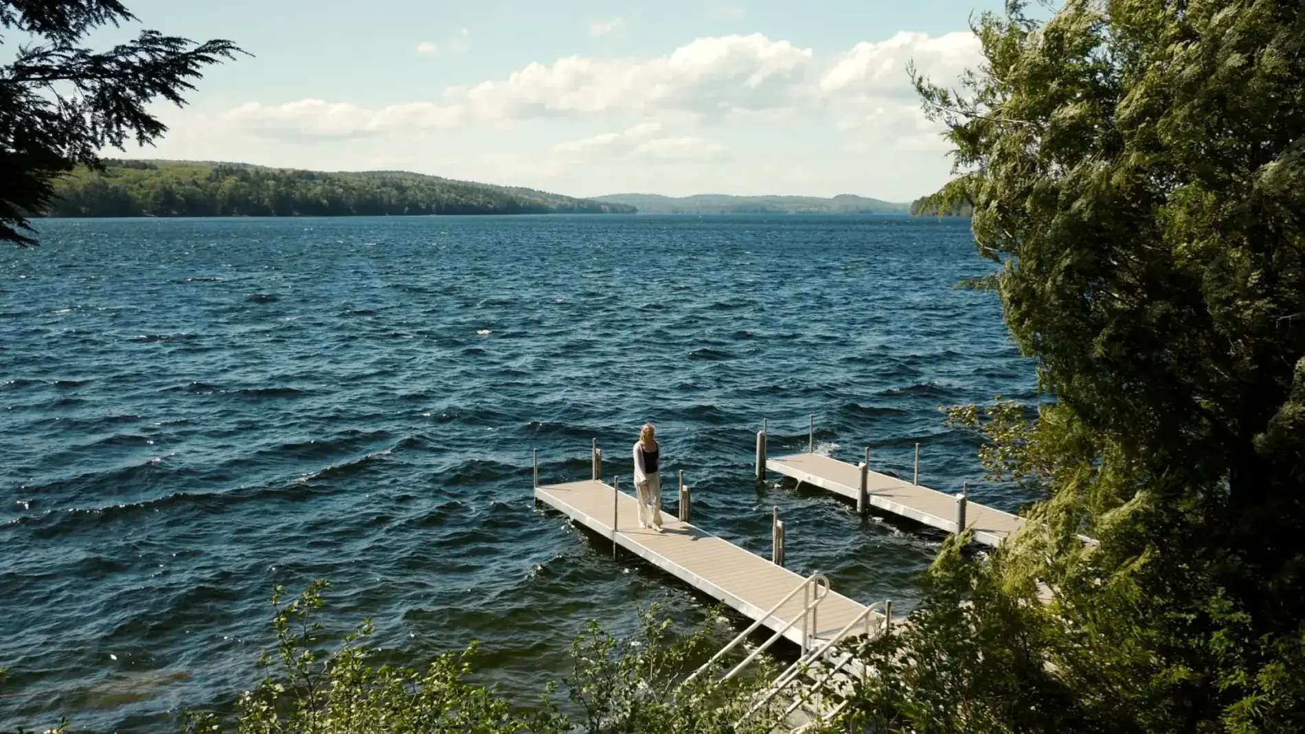 A person stands on a wooden dock extending into a large, rippling lake, surrounded by trees, with hills visible in the distance under a partly cloudy sky.
