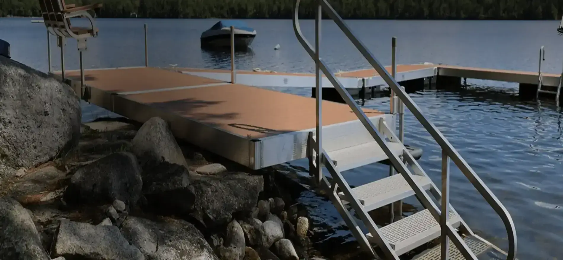 A dock with metal stairs leads from rocky shore into a calm lake. A boat floats nearby, and trees line the far shore under a clear sky.